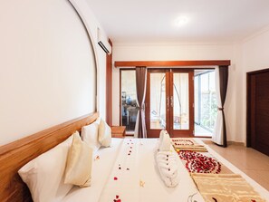 A hotel room with a bed, a wooden headboard, a large window with curtains, and a view of the outside. - Amaluku Villa (Ubud)