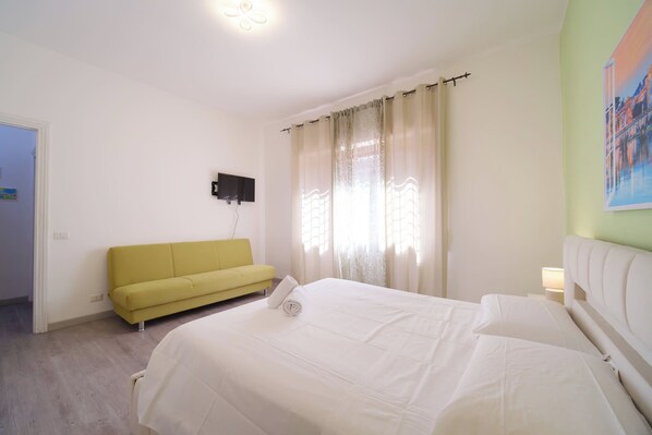 A bedroom with a bed, a sofa, a TV mounted on the wall, and a window with curtains.