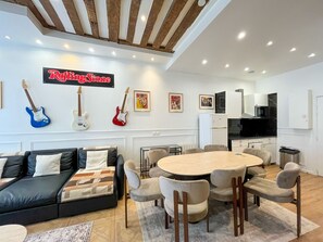 A modern living room with a large sofa, a dining table, and chairs. There are guitars on the wall, a kitchen area, and a sign that reads 'RollingStone'.