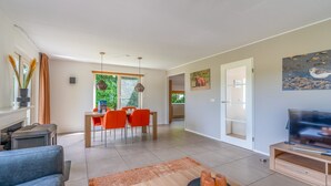 A modern living room with a sofa, a dining area with orange chairs, a TV, and artwork on the walls. - Charming Villa With Sauna on the Water (De Koog)