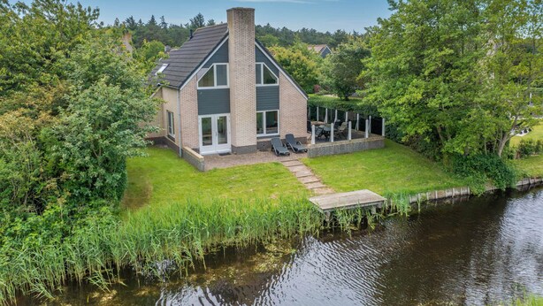 A modern house with a large deck, a pond, and lush greenery. - Charming Villa With Sauna on the Water (De Koog)