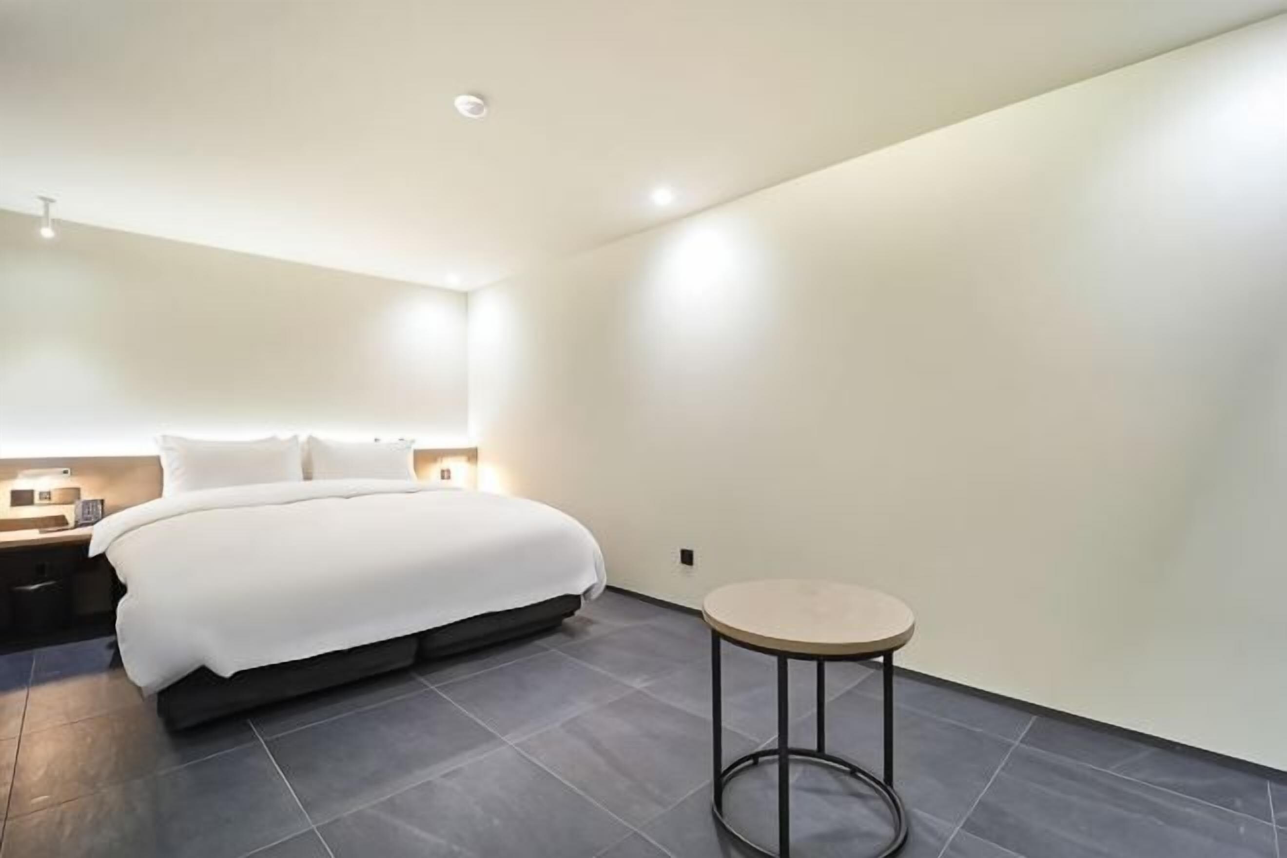 Moderni hotellihuone, jossa on suuri sänky, yöpöydät, pyöreä pöytä ja pelkistetty sisustus.