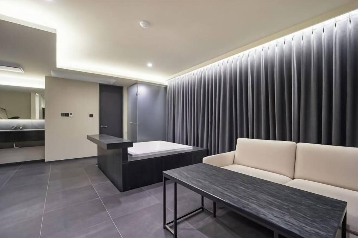 Una habitación de hotel moderna con sofá, una mesita, un banco integrado, un espejo grande y una pared cubierta de cortinas.