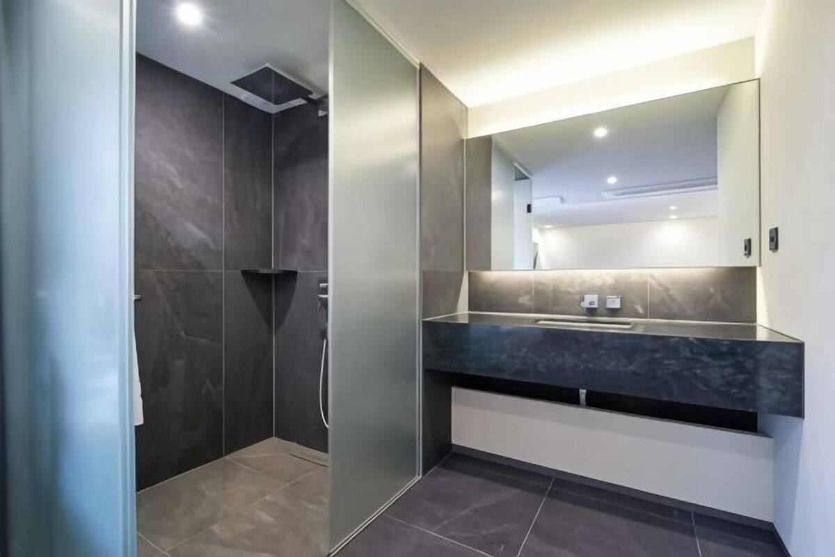 Un baño moderno con ducha de baldosas oscuras, un espejo grande y un lavamanos sobre encimera oscura.