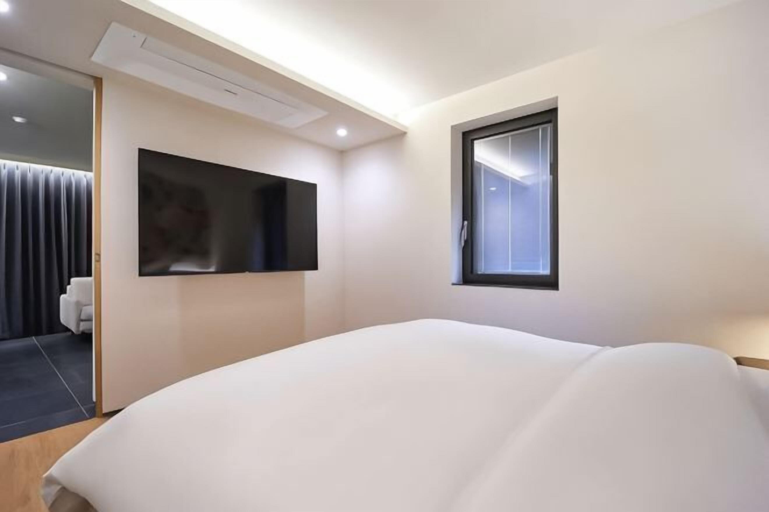 Moderni hotellihuone, jossa on suuri sänky, seinälle asennettu televisio, ikkuna ja ovi, joka johtaa kylpyhuoneeseen.