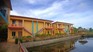 A row of yellow buildings with balconies, a canal in front, and a small hut in the background. - CHUTI BEACH REOSORT (Mandarmani)