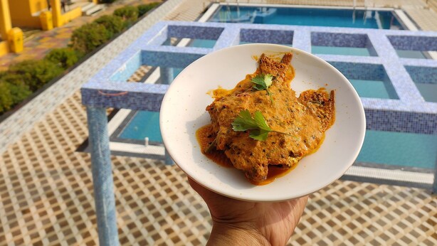 A hand holding a plate of food with a pool and buildings in the background. - CHUTI BEACH REOSORT (Mandarmani)
