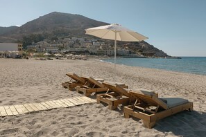 Una playa con sillones de madera y una sombrilla grande, un paseo marítimo de madera y edificios al fondo.