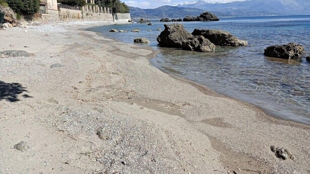 Uma praia com águas cristalinas, afloramentos rochosos e um edifício com fachada clara.