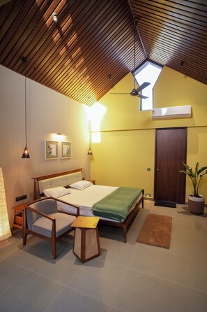 A bedroom with a sloped ceiling, a bed, a chair, a small table, and a potted plant. - Kshitij Villa (Bhuj)