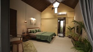 A bedroom with a bed, bedside tables, a desk, a chair, a green rug, and a ceiling fan. - Kshitij Villa (Bhuj)