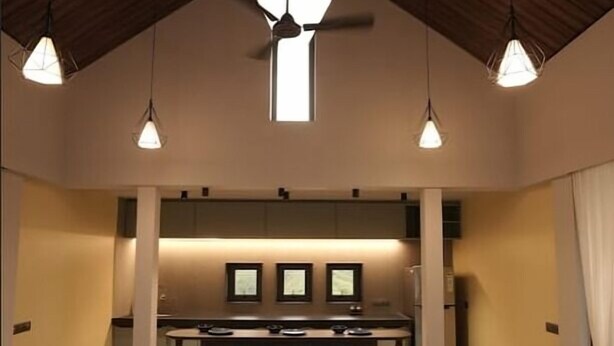 A modern kitchen with a central island, pendant lighting, and a high ceiling with exposed beams. - Kshitij Villa (Bhuj)