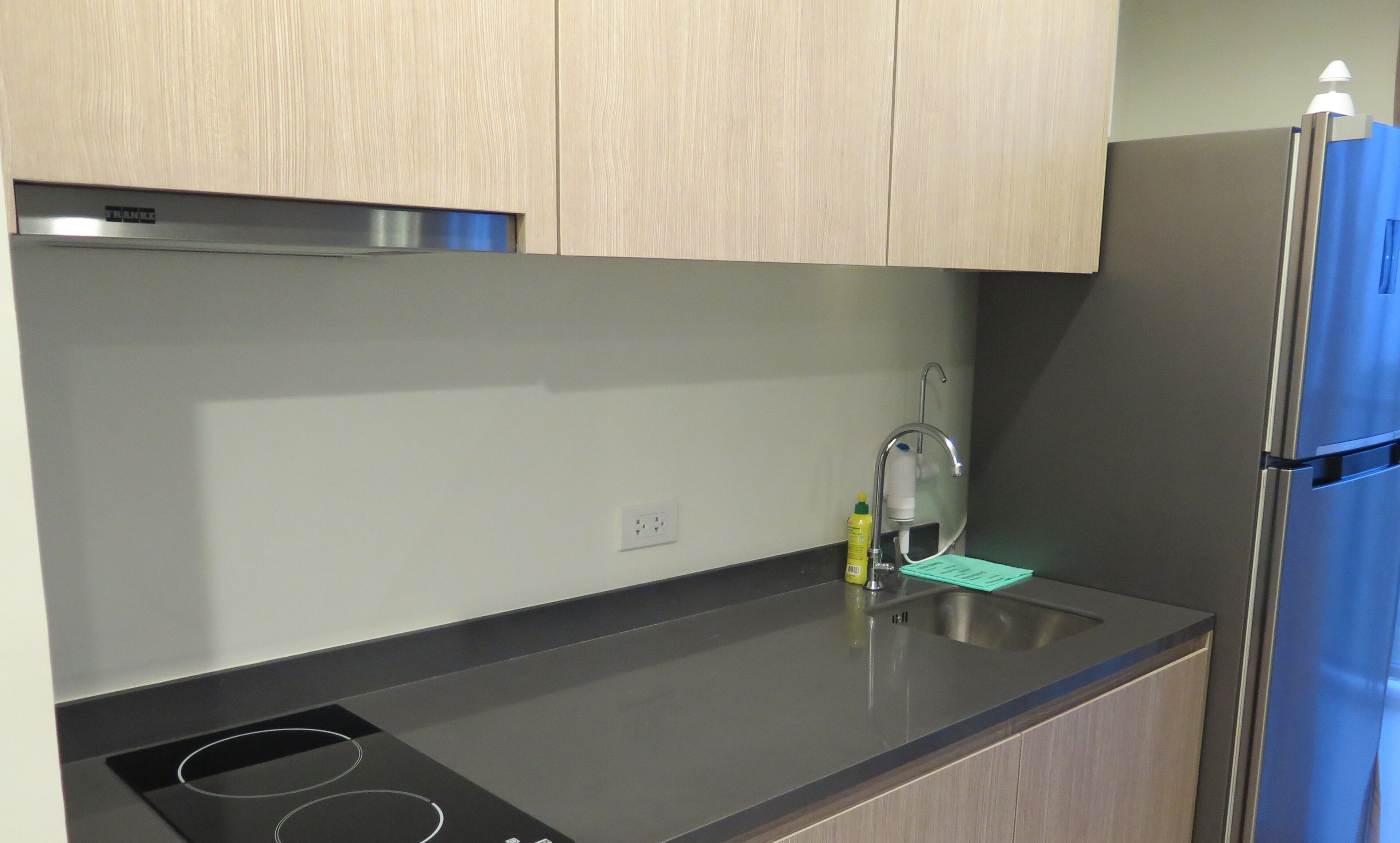 A modern kitchen with light wood cabinets, a stainless steel refrigerator, and a black countertop.