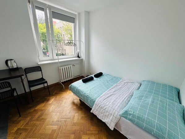 A small, tidy room with a wooden floor, a bed with a blue and white bedspread, a radiator, a window with a view of buildings, and a desk with a kettle and chair.
