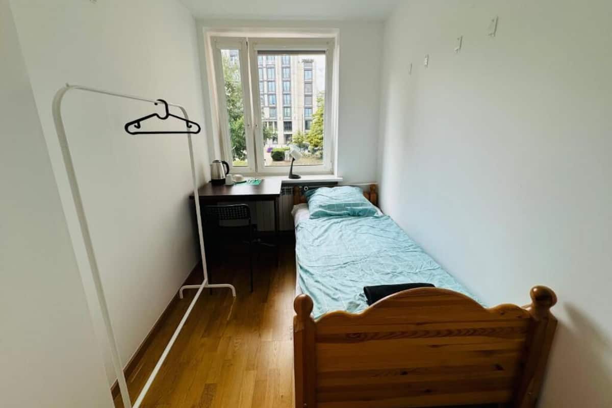 A small, bright room with a wooden bed, a desk with a chair, and a window with a view of buildings and trees.