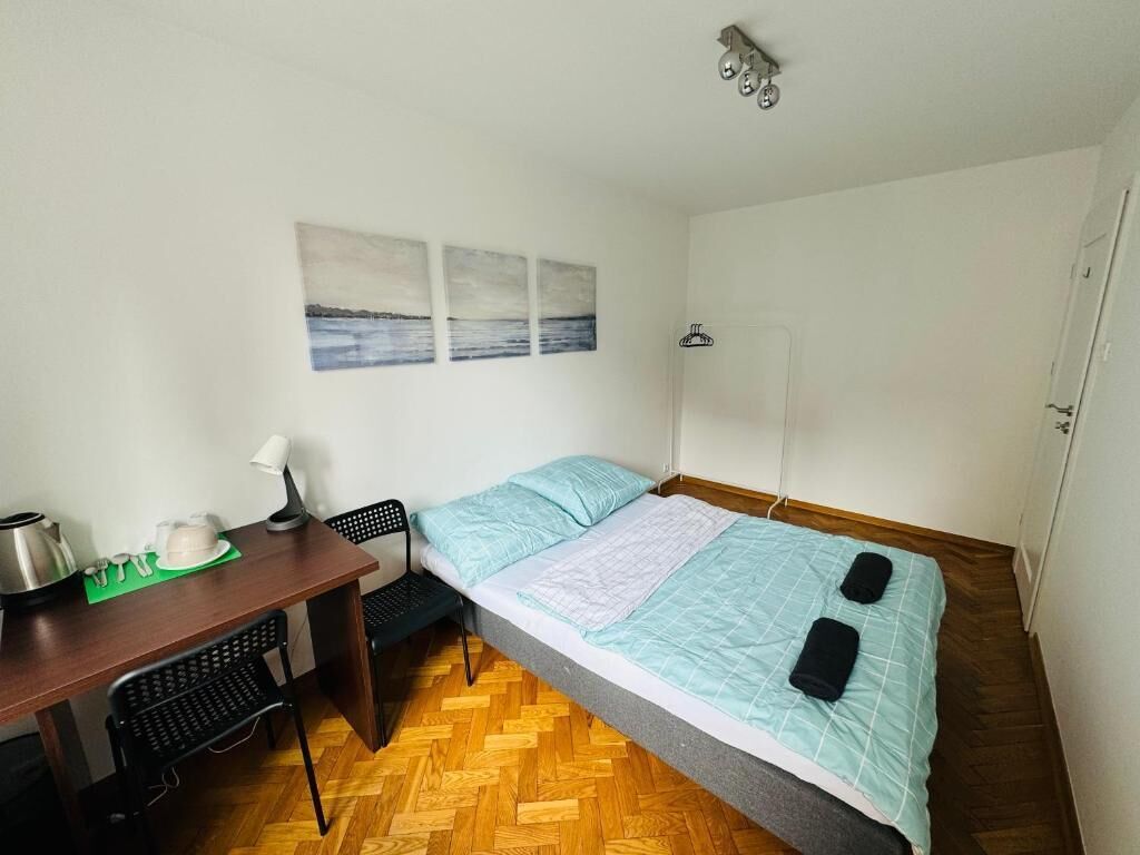 A small, neatly arranged room with a bed, a desk, and a chair. There are wall-mounted artworks above the bed and a shower area to the right.