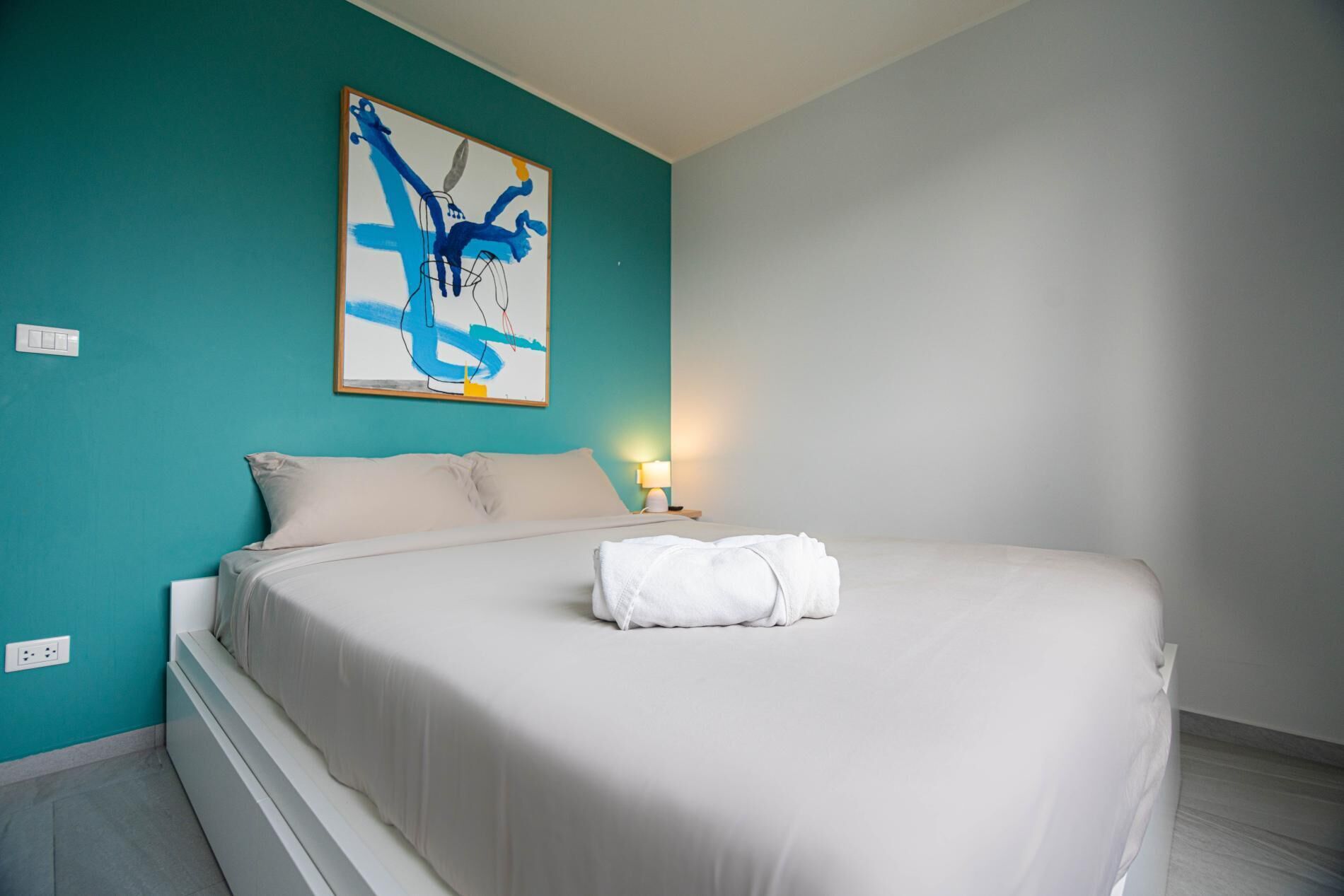 A bedroom with a bed, a wall-mounted abstract painting, a bedside lamp, and a power outlet.