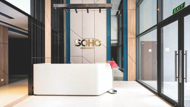 A modern reception area with a white reception desk, a sign reading "SOHO RESIDENCE," and an exit sign above a glass door. - The Heart of Saigon Soho Central (Ho Chi Minh City)