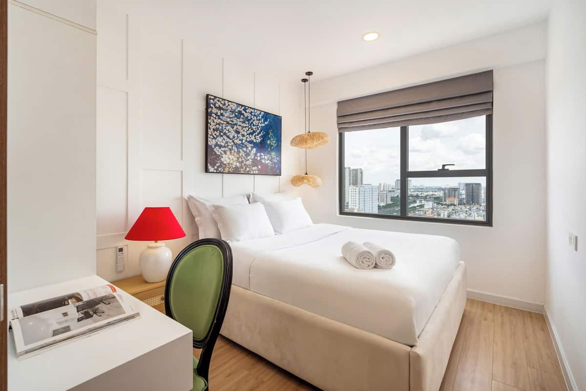 A hotel room with a bed, a desk, a chair, a red lamp, a city view, and a painting on the wall.