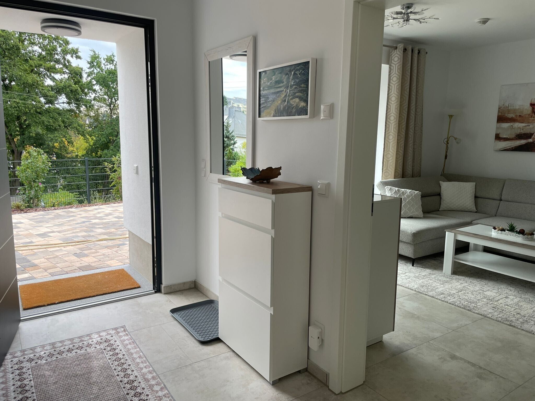 A modern living room with a sofa, a coffee table, and a dining table. There is a door leading to a balcony with a view of trees and a fence.