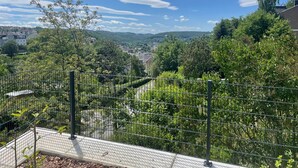 Une vue depuis un balcon doté d’une rambarde en métal, donnant sur un paysage verdoyant et luxuriant, avec des arbres et une ville située au loin.
