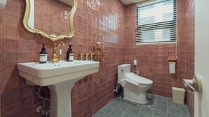Un baño con lavamanos blanco, grifos dorados, inodoro y ventana con persianas.