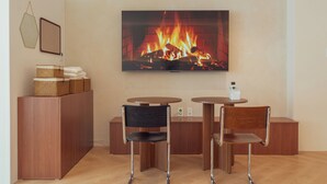 Una sala moderna con un televisor de pantalla plana mostrando una chimenea, dos sillas de madera, una mesita pequeña y un mueble de madera con cestas.