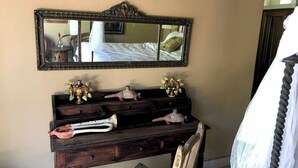 Une commode en bois avec un miroir au-dessus, une petite table ornée de quelques objets décoratifs et une chaise recouverte d’une housse en dentelle blanche.