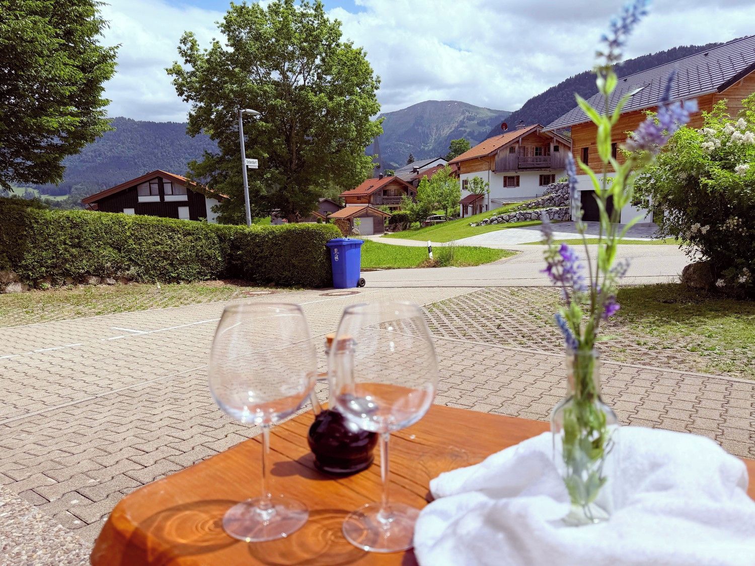 Une table avec des verres à vin et un vase de fleurs, placée sur un chemin pavé, d’où l’on peut admirer des maisons et des montagnes.