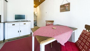 Et lille, hyggeligt rum med trælofter, et hvidt skab med skuffer, et fjernsyn ovenpå, et bord med rød-hvidternet dug og en stol med rød pude.