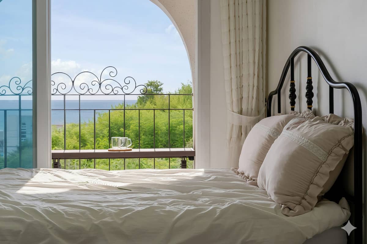 A bedroom with a large window, a bed with pillows, and a view of greenery and a body of water.