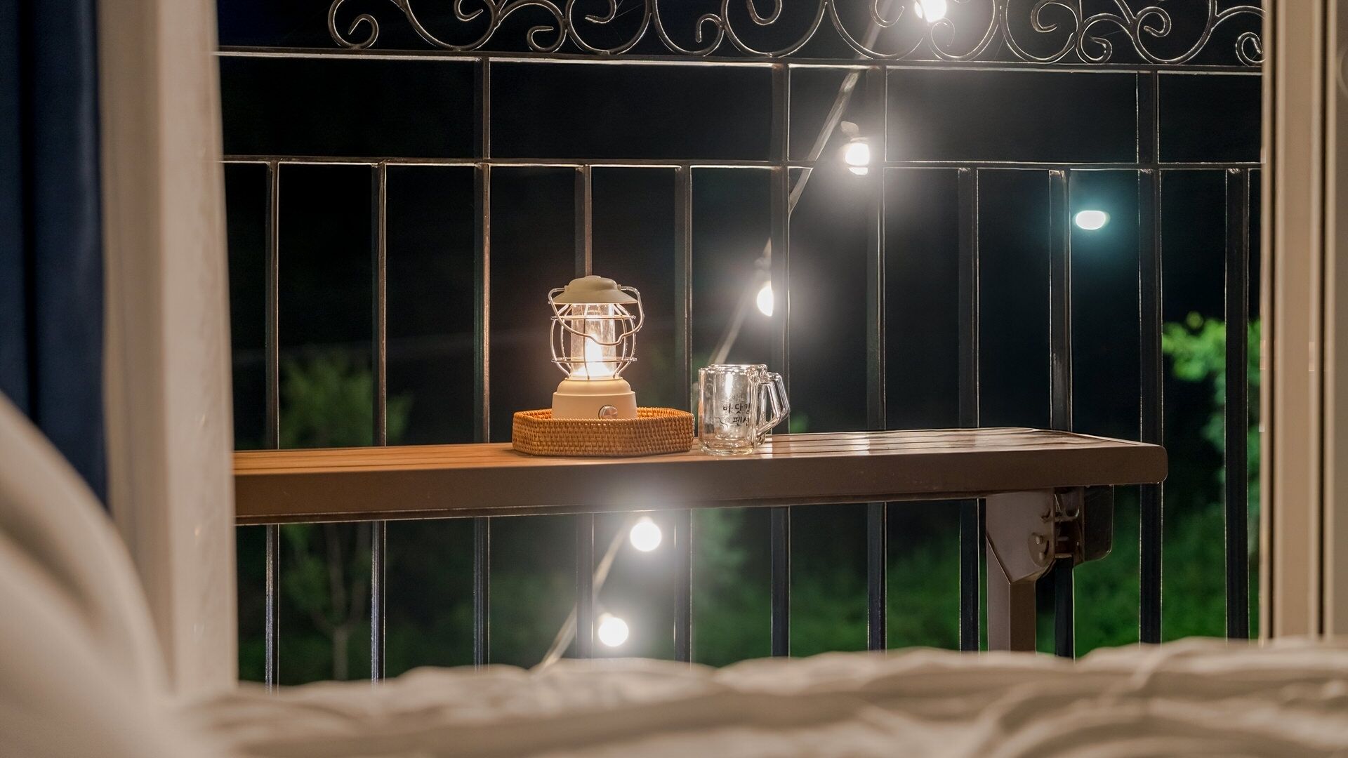 A window with a decorative metal grille, a white lamp, and a clear glass mug on a wooden ledge.