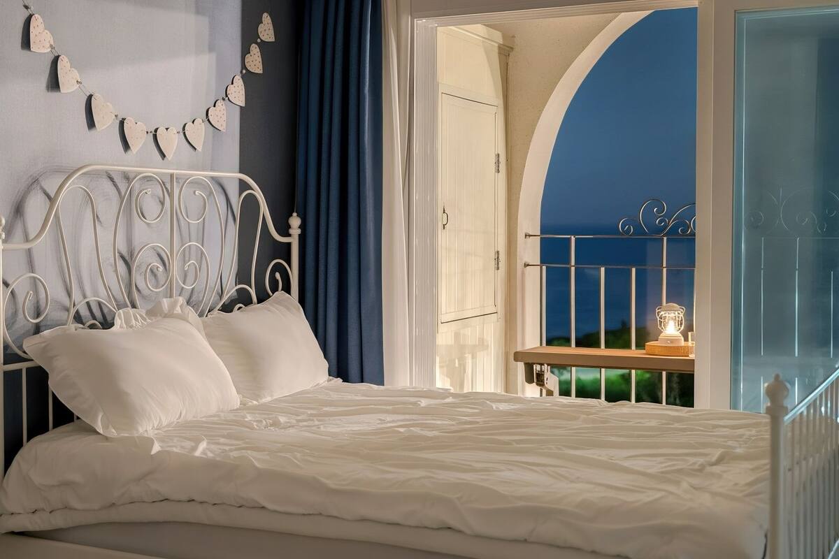A bedroom with a white metal bed frame, white bedding, a wall-mounted heart decoration, and a view of the night sky through an arched window.