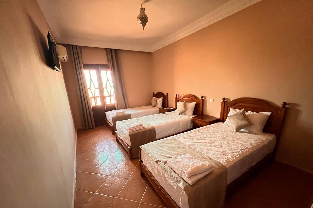 A hotel room with two single beds, a wooden headboard, a small bedside table, a nightstand, and a window with curtains.
