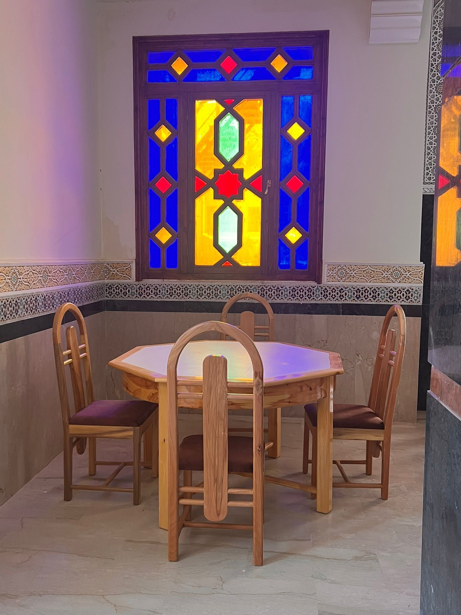Een eetgedeelte met een ronde houten tafel en vier stoelen, een decoratief glas-in-loodraam erboven en een patroon langs de muur.