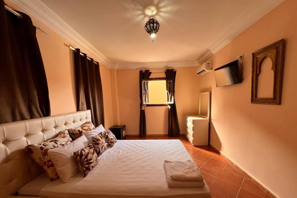 A hotel room with a large bed, a bedside table, a TV mounted on the wall, and a window with curtains.