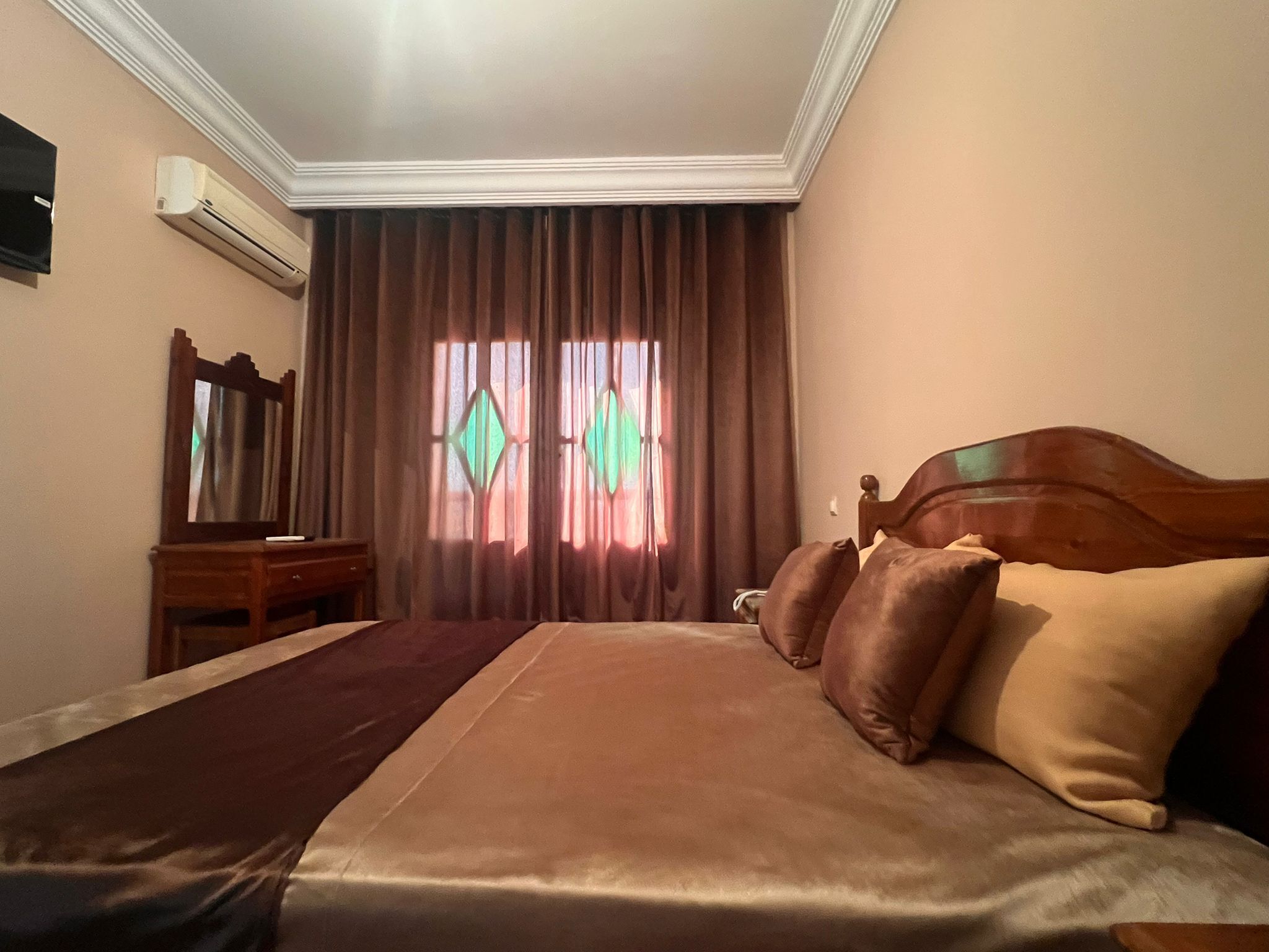 Une chambre d’hôtel avec un grand lit, une tête de lit en bois, une table de chevet, un miroir et une fenêtre avec des rideaux.