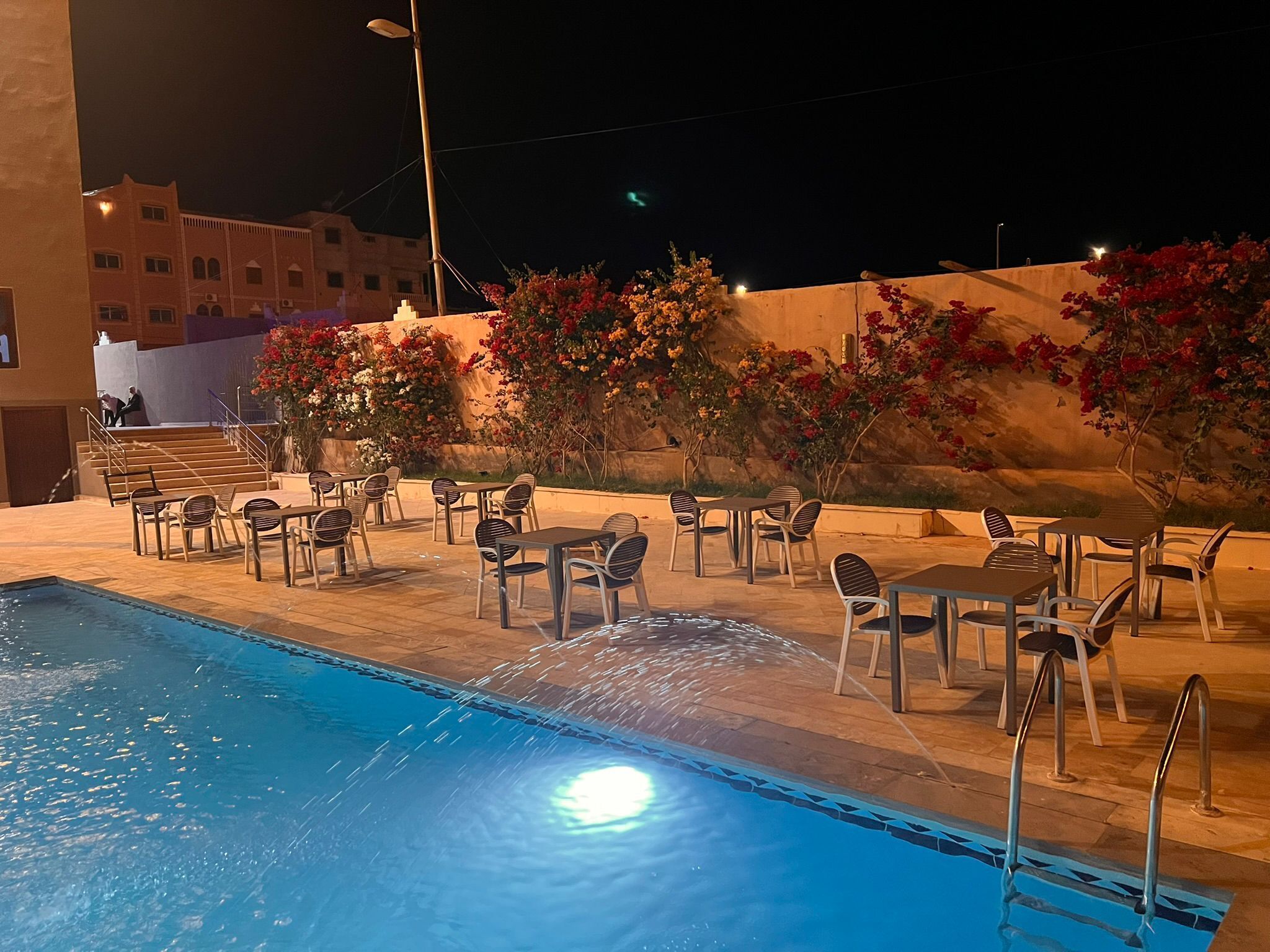 Un espace aménagé au bord de la piscine, avec des chaises et des tables, une piscine et un bâtiment en arrière-plan.