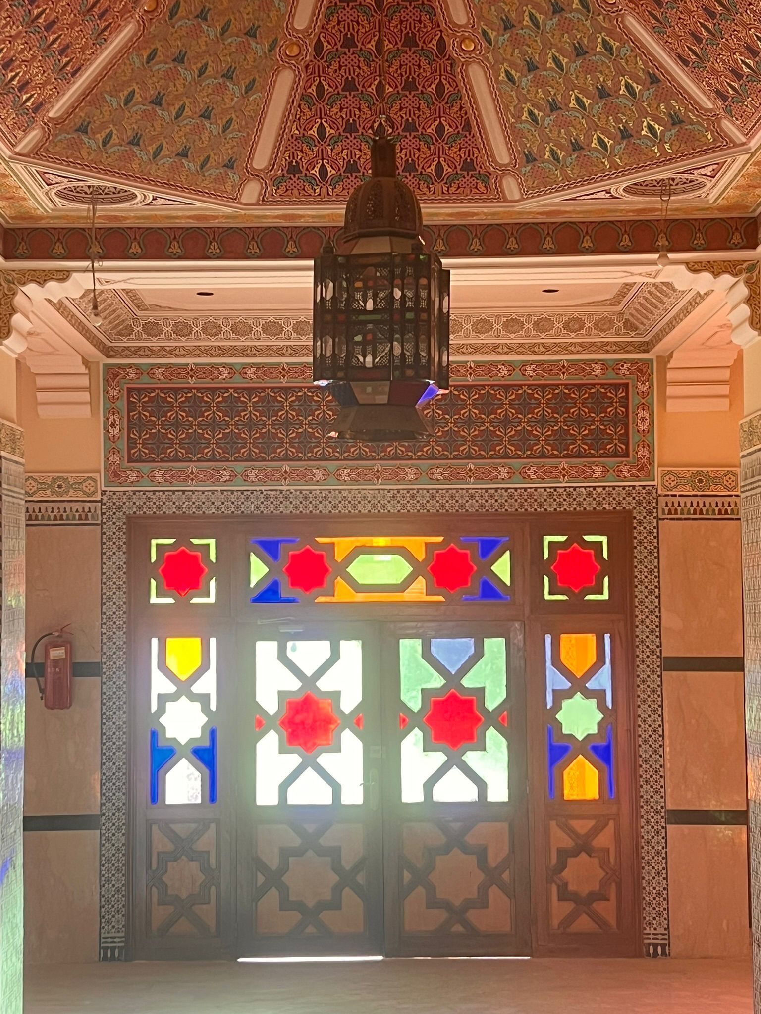 Un interior ornamentado con un techo detallado, un farol central y una puerta con diseños geométricos.