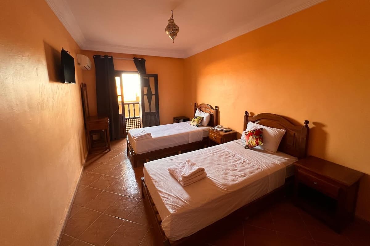 A room with two single beds, a wooden nightstand, a small table, and a ceiling light fixture.