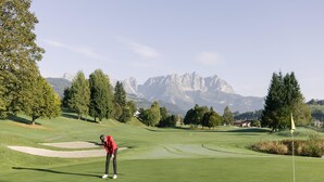 En golfbana med en golfspelare som förbereder sig för att slå ett putt, omgiven av träd och med berg i bakgrunden.