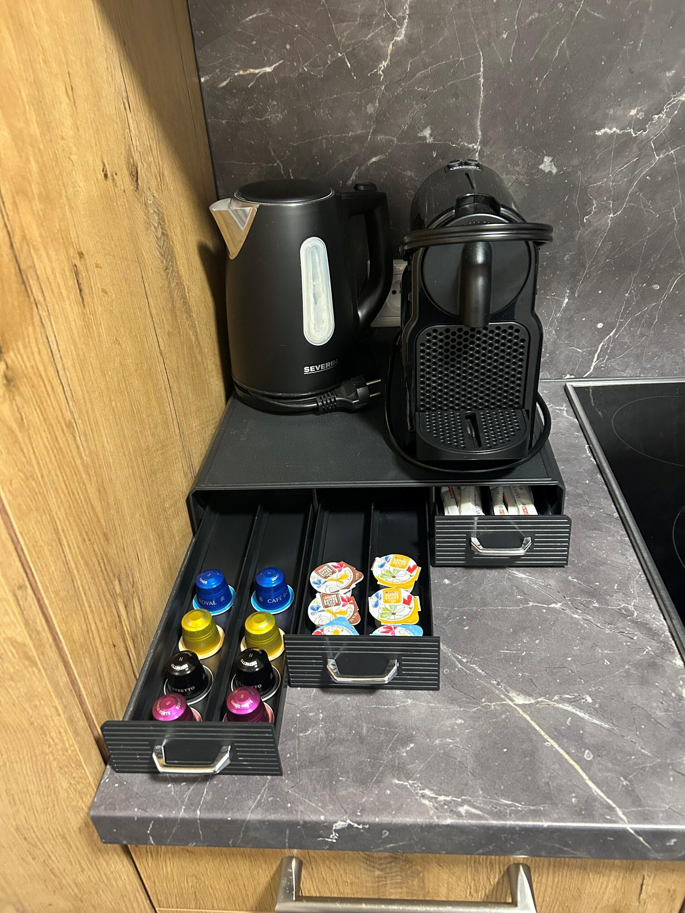 Un plan de travail de cuisine avec une machine à café, une bouilloire et un tiroir contenant des dosettes de thé et de café.