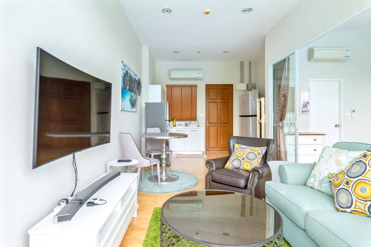 Een moderne woonkamer met een flatscreen televisie, een bank, een salontafel, en een eethoek met een kleine tafel en stoelen.