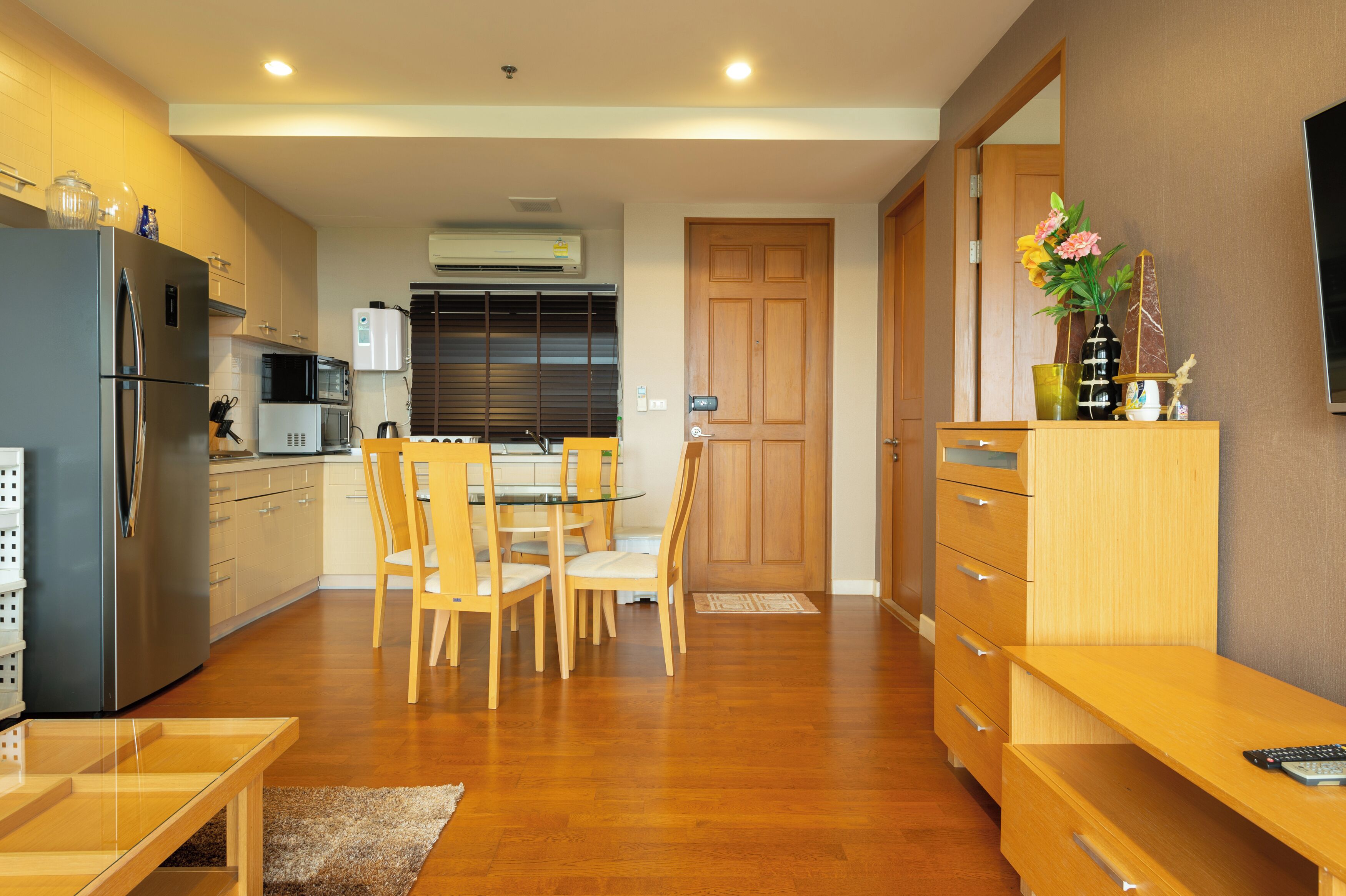 A modern kitchen with stainless steel appliances, a dining area with wooden chairs and a glass table, and a wooden cabinet with a vase of flowers.