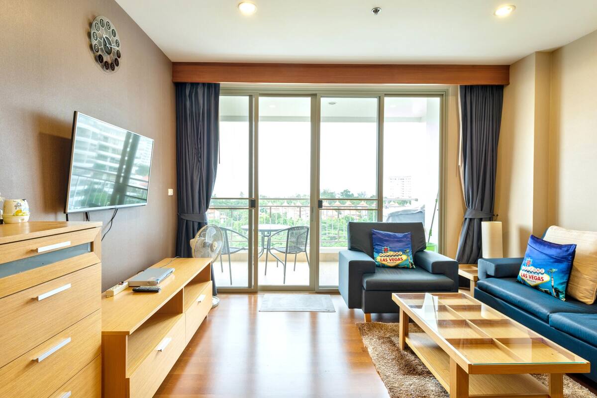 Een moderne woonkamer met een flatscreen-tv, houten meubels, een bank met decoratieve kussens en een balkon met uitzicht.