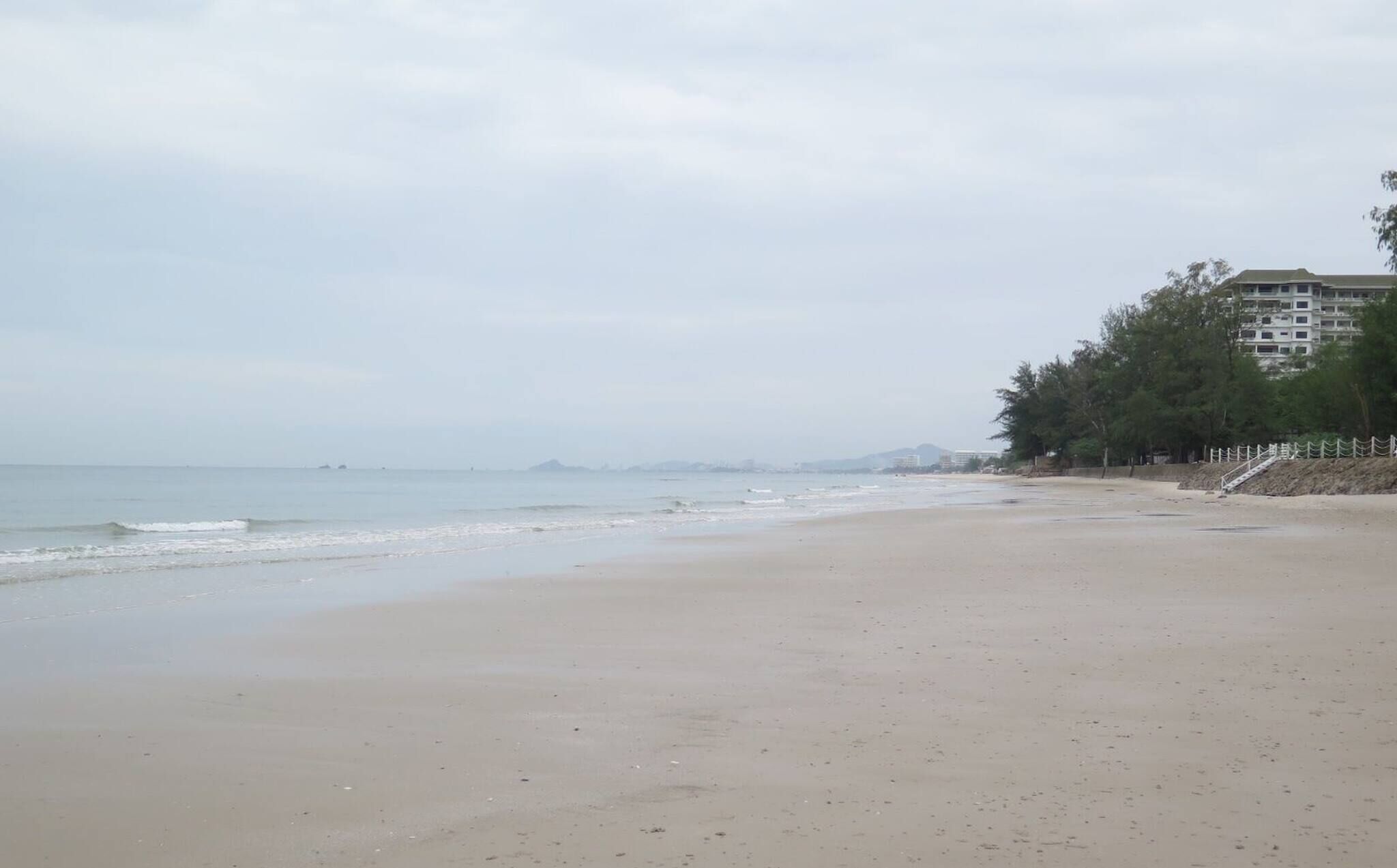A wide sandy beach with gentle waves, a clear sky, and a building with a fence on the right.
