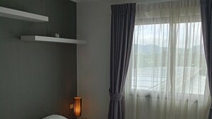 Una camera d'albergo con un letto, un comodino con una lampada, uno scaffale sopra il letto e una finestra con tende.