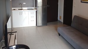 Una cucina moderna con mobili bianchi, un frigorifero in acciaio inossidabile e un forno integrato. C'è un divano grigio e un piccolo tavolo da pranzo con una sedia.
