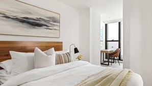 Een moderne slaapkamer met een groot bed, een houten hoofdbord, een nachtkastje met een lamp, en een stadszicht door grote ramen.