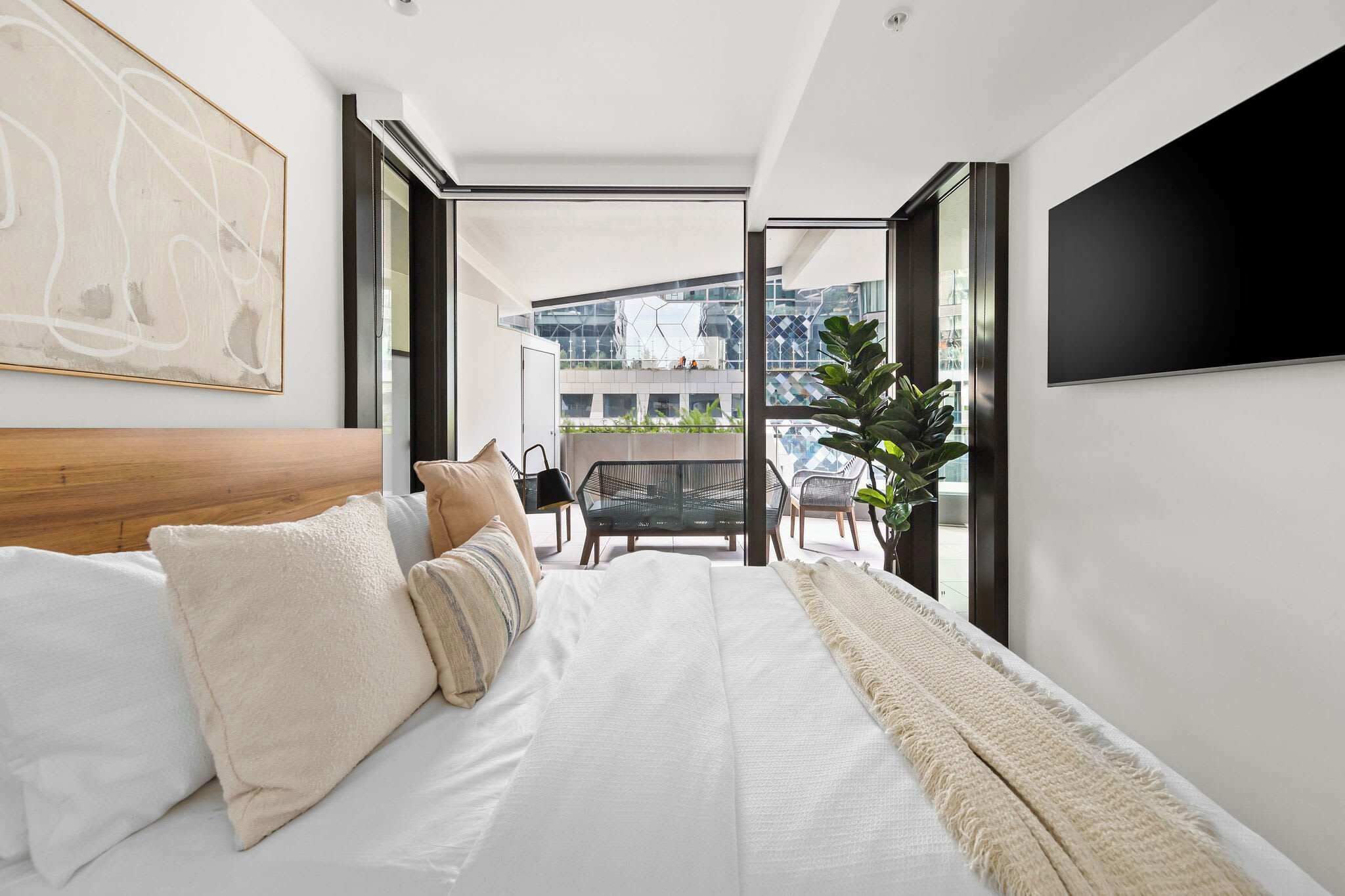 A modern bedroom with a large bed, a view of the city through a glass door, and a mounted TV on the wall.